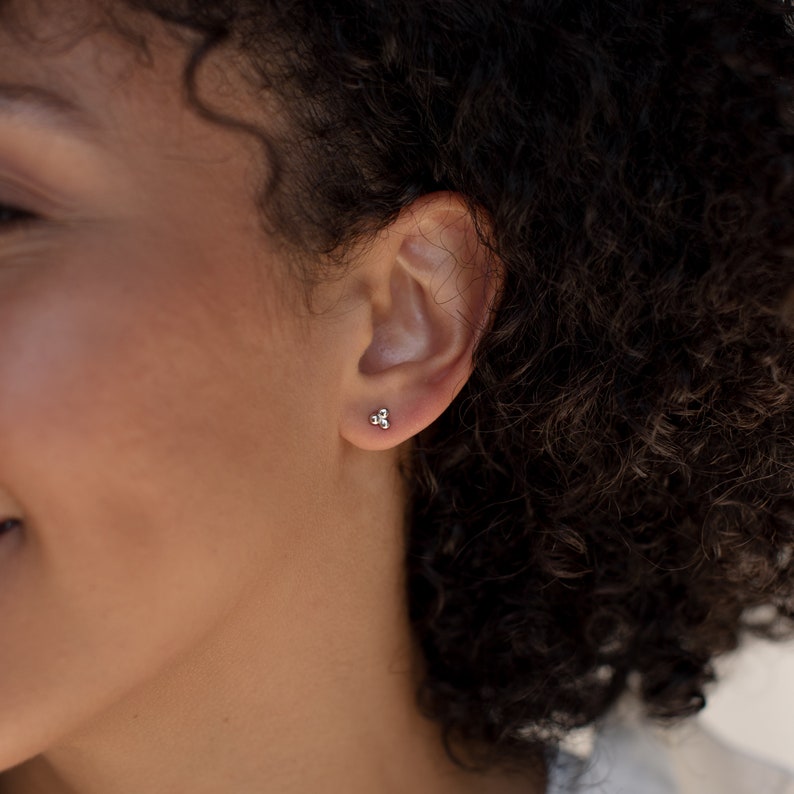 May include: A close-up of a person's ear with a small silver stud earring featuring three tiny balls.