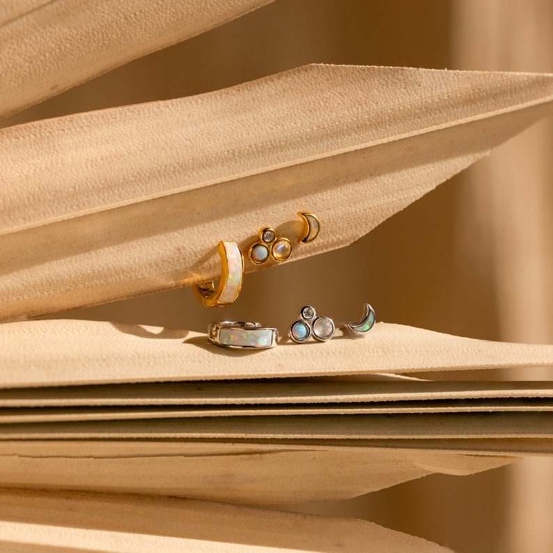 May include: A set of three opal and moon stud earrings in gold and silver. The earrings are arranged on a beige background.