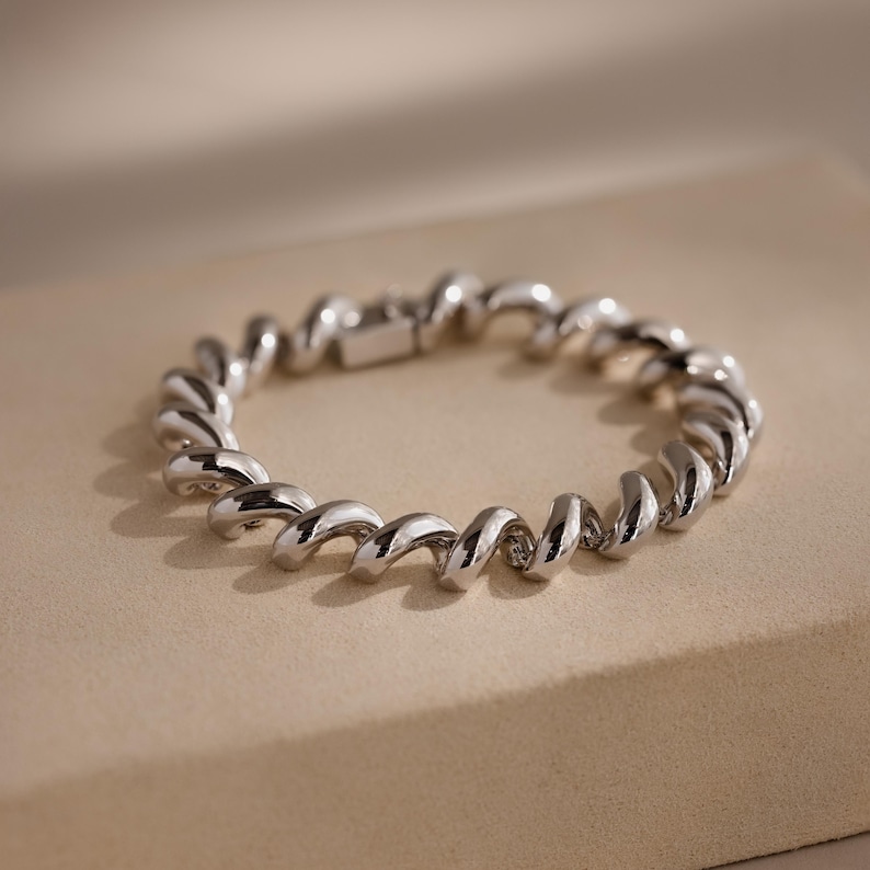 May include: A silver bracelet with a spiral design. The polished metal bracelet has a clasp closure. The design is a continuous, twisting pattern, creating a modern and elegant look. The bracelet is displayed on a neutral background.