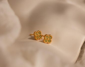May include: A pair of gold stud earrings featuring a four-leaf clover shape. Each earring is crafted with a metallic gold frame and contains four small, round, light green stones arranged in a cluster to resemble a clover leaf design.