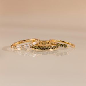 May include: Three gold rings with different designs. One ring features clear baguette and round-cut stones. Another has olive-green square stones. The third ring has round olive-green stones and clear stones. All rings are on a reflective surface.