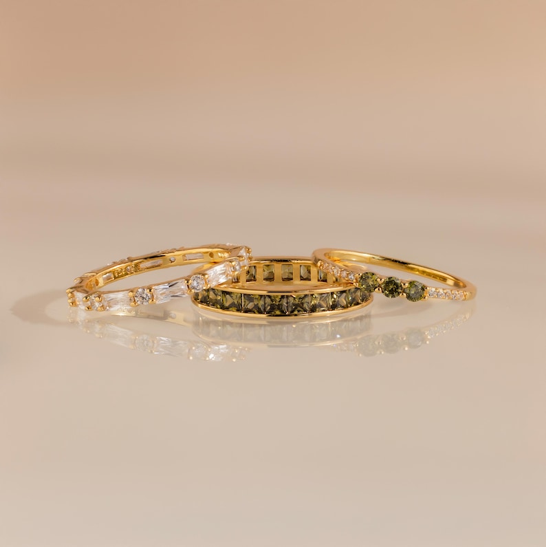 May include: Three gold rings with different designs. One ring features clear baguette and round-cut stones. Another has olive-green square stones. The third ring has round olive-green stones and clear stones. All rings are on a reflective surface.