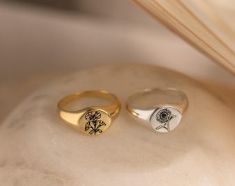Puede incluir: Dos anillos tipo sello, uno dorado y otro plateado, cada uno con una cara circular plana. El anillo dorado está grabado con un diseño floral negro, mientras que el anillo plateado muestra una ilustración de un girasol negro en su superficie.