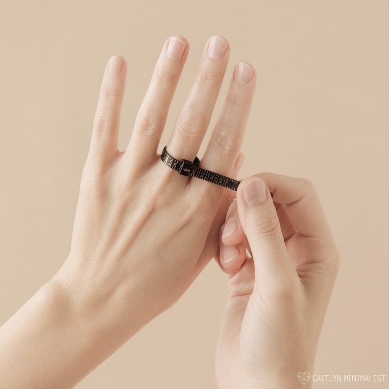May include: A black ring sizer is wrapped around a finger to measure its size. The sizer is a flexible band with measurement markings. The background is a solid, neutral colour. The image is well-lit and focuses on the ring sizer.
