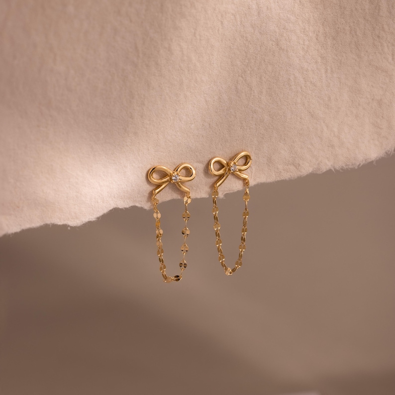 May include: A pair of gold-colored bow earrings with a small diamond in the center of each bow. The bows are connected to a delicate chain of small, flat, gold-colored links.