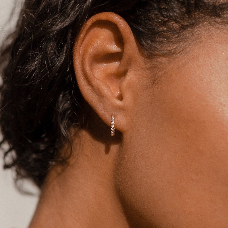 May include: A gold hoop earring with small, clear gemstones. The earring is worn on the ear of a person with dark hair and skin. The earring is a simple, elegant design.