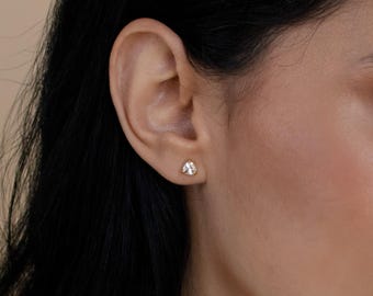 Op de afbeelding: Een kleine, driehoekige oorsteker met een heldere, gefacetteerde edelsteen in een goudkleurige metalen zetting, stevig geplaatst in de oorlel. De oorbel heeft een gepolijste afwerking en een eenvoudig, geometrisch ontwerp dat plat tegen de huid aanligt.