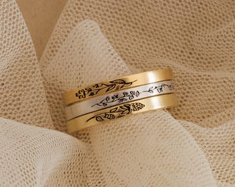 May include: A set of three stacked rings featuring two gold-toned bands and one silver-toned band. Each ring is engraved with a distinct black floral botanical design, showcasing delicate stems, leaves, and small flower buds in a linear pattern.