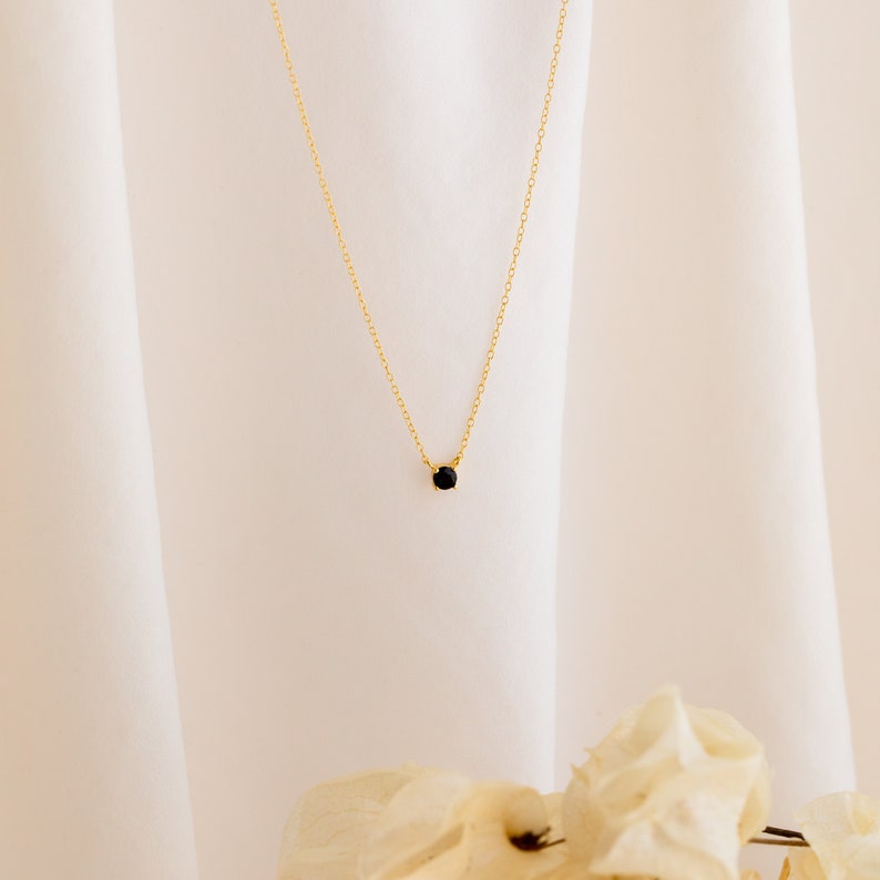 A close up of our Onyx Dainty Charm Necklace in 18K Gold finish hanging against an off-white fabric background with cream flowers in the foreground - featuring a small round 4mm black CZ Diamond Charm on a dainty cable chain.