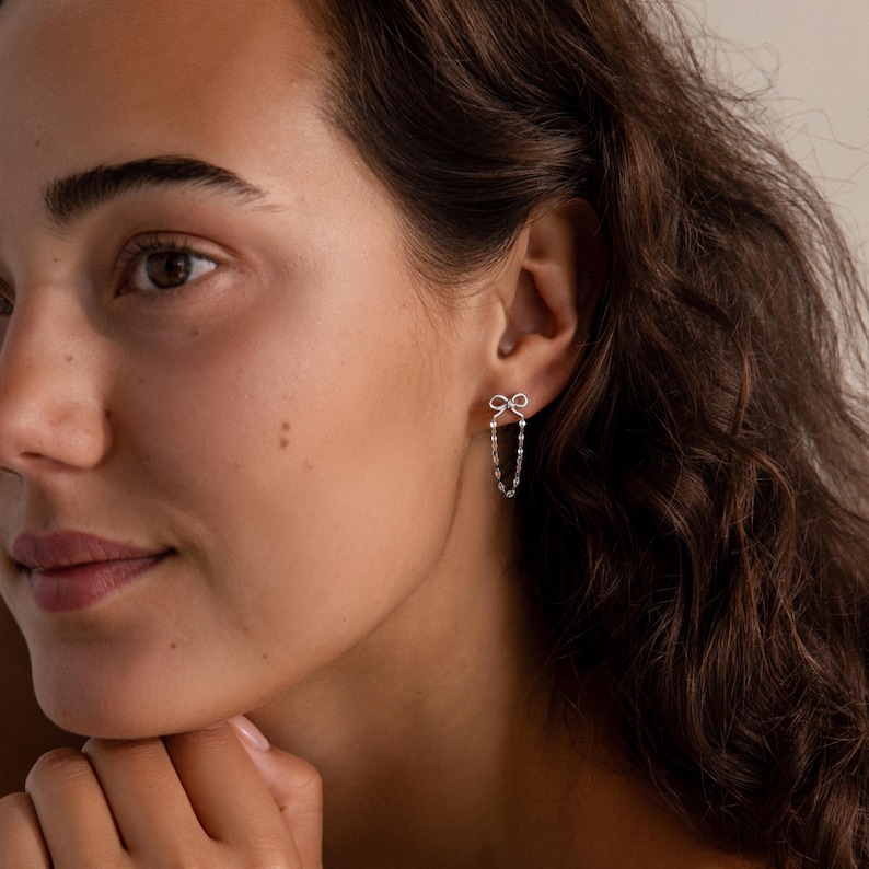 Woman wearing Bow Chain Stud Earrings in silver with delicate bow studs and draped chain loops, styled with natural makeup and wavy hair.