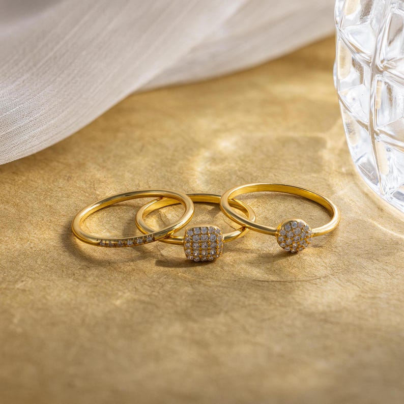May include: Three gold rings with small, square-shaped stones set in the center. The stones are covered in tiny, sparkling diamonds.
