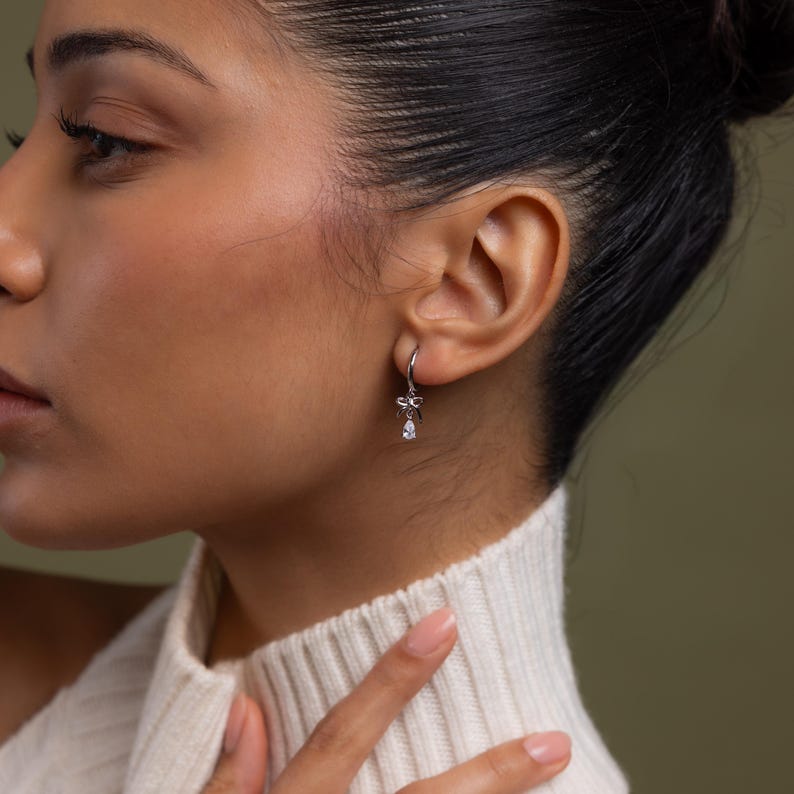 May include: A close-up of a silver earring with a bow design and a dangling teardrop-shaped clear stone. The earring is attached to a silver hoop. The person is wearing a white sweater.