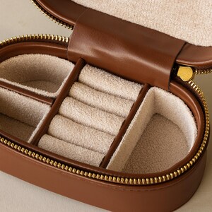 May include: A brown leather jewelry box with a gold zipper. The inside of the box is lined with a light brown fabric and has multiple compartments for storing rings and other jewelry.