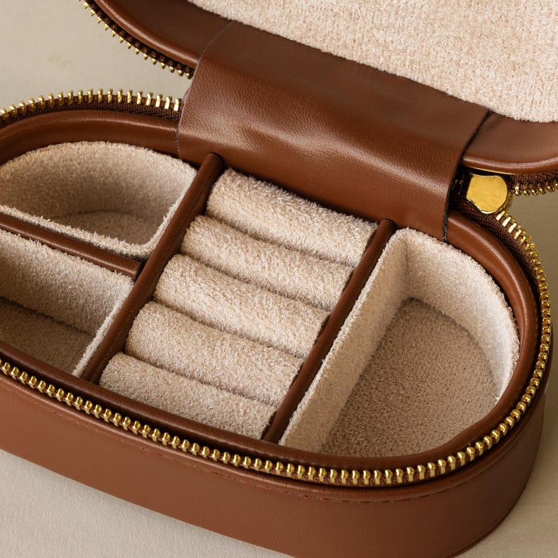 May include: A brown leather jewelry box with a gold zipper. The inside of the box is lined with a light brown fabric and has multiple compartments for storing rings and other jewelry.