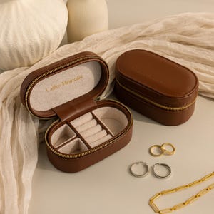 May include: Two brown oval shaped leather jewelry boxes with gold zippers. The box on the left is open and has a white interior with compartments for rings and earrings. The box on the right is closed and has the text 'Carry Me With You' embossed on the top. The boxes are on a white fabric surface with gold hoop earrings, a gold chain necklace, and a silver hoop earring.
