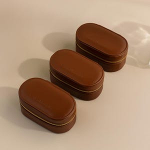 May include: Three brown oval-shaped leather jewelry boxes with gold zippers. Each box has the words 'Colibri Jewelry' embossed on the top.