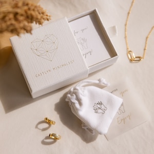 K&ouml;nnte beinhalten: Eine wei&szlig;e Box mit goldener Beschriftung "Caitlynn Minimalist" und einem geometrischen Herzdesign. Die Box enth&auml;lt einen kleinen wei&szlig;en Beutel mit goldener Beschriftung "Crafted for you" und eine goldene Kettenkette mit einem Herzanh&auml;nger.