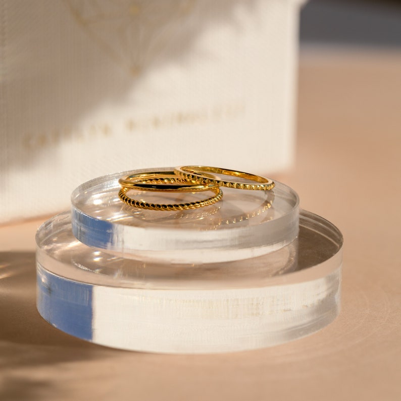 A close up of our Dainty Stacking Ring Set in 18K Gold finish placed on a clear decorative dish with our Caitlynminimalist gift box in the background - featuring a set of 3 rings: 1 Thin Band, 1 Twist Ring and 1 Notched Ring all about 1mm thick.