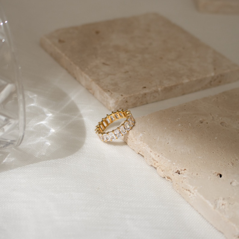 May include: A gold ring with a row of clear rectangular stones. The ring is on a light brown textured surface.