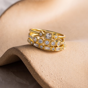 May include: A stack of four gold-colored rings with various gemstone settings. The rings feature round, square, and baguette-cut clear gemstones. The rings are displayed on a textured, light brown surface.