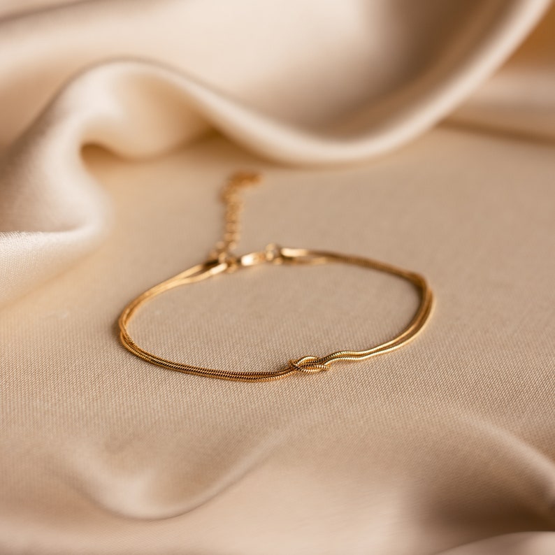 May include: A delicate gold chain bracelet with a knot design. The bracelet is on a light beige fabric background.