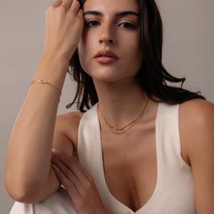 May include: A gold-toned necklace and bracelet set. The necklace features two delicate chains, and the bracelet has a knot design. The jewelry is worn on a person wearing a white tank top.