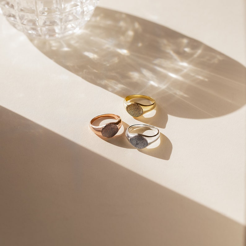 A close up of 3 of our Fingerprint Signet Rings in 18K Gold, Sterling Silver, and Rose Gold finish placed on a plain white table with soft lighting - each ring features an 8mm Signet Design engraved with a Custom Fingerprint Design.