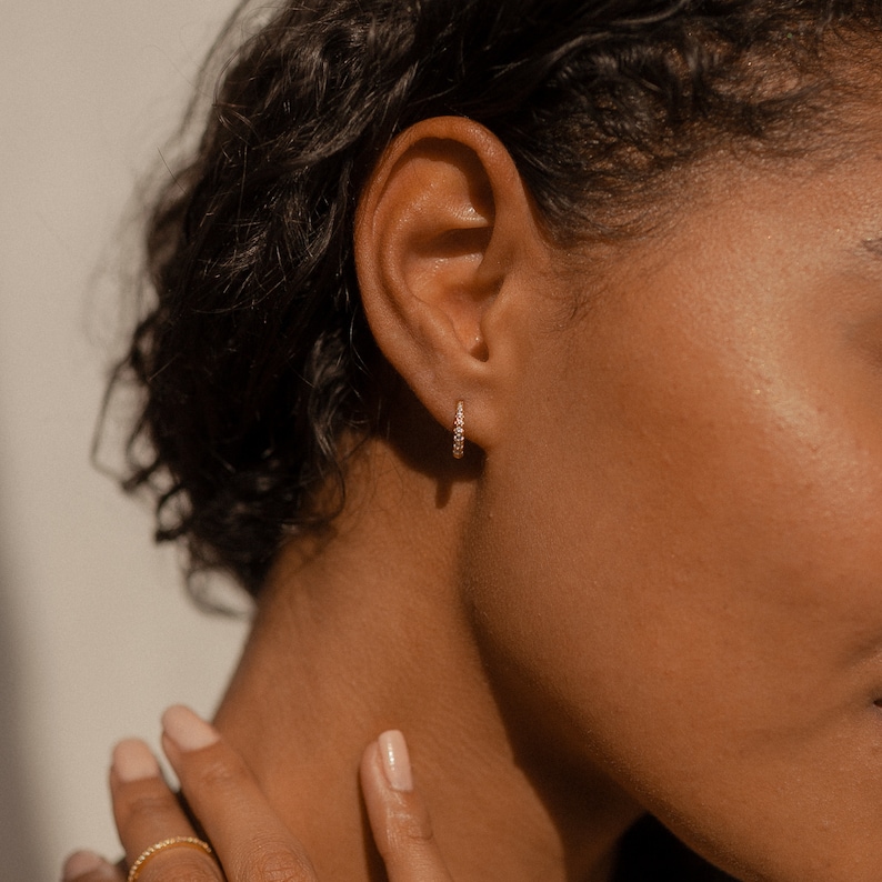 May include: A close-up shot of a person's ear adorned with a small, gold hoop earring. The earring is encrusted with small, sparkling stones. The person's skin tone is a warm brown, and their hair is dark and curly.