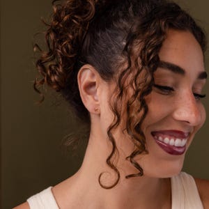 Gold letter K initial stud earring worn on the ear, shown on a model with curly hair styled up, highlighting the small polished letter design against the skin.