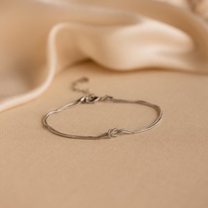 May include: A silver chain bracelet with a knot design. The bracelet is on a light brown surface.