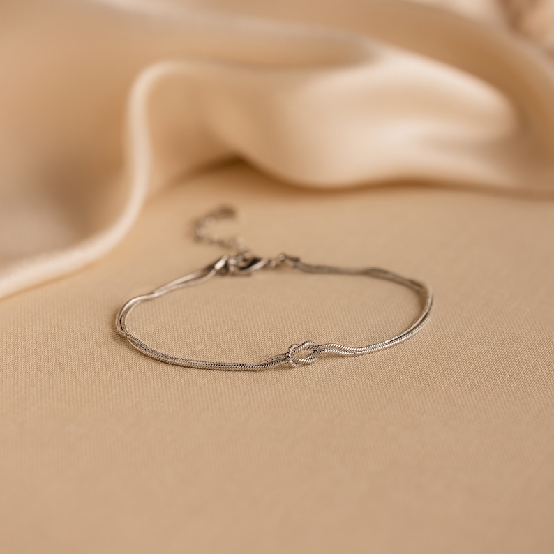 May include: A silver chain bracelet with a knot design. The bracelet is on a light brown surface.