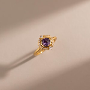 May include: A gold ring featuring a central hexagonal purple gemstone set in a bezel. The centre stone is surrounded by a halo of small, clear, round-cut white stones, with two larger white stones positioned at the top and bottom of the setting.