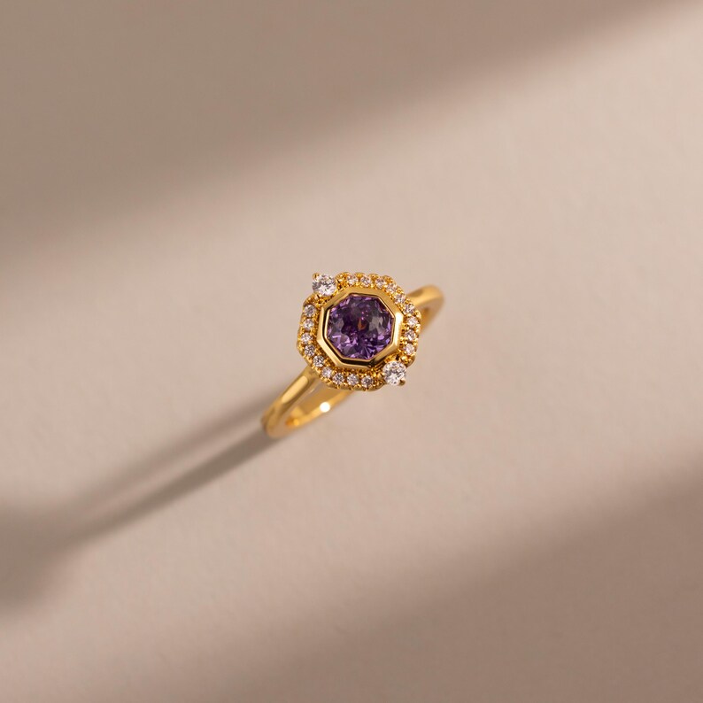 May include: A gold ring featuring a central hexagonal purple gemstone set in a bezel. The centre stone is surrounded by a halo of small, clear, round-cut white stones, with two larger white stones positioned at the top and bottom of the setting.