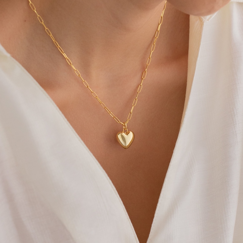 Gold chain necklace with elongated links and a polished heart shaped pendant, styled with a white button-down top.
