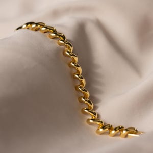 May include: A gold bracelet with a spiral design. The bracelet is made of linked, curved segments, creating a flowing, elegant look. The jewellery is displayed on a cream-coloured, textured fabric.