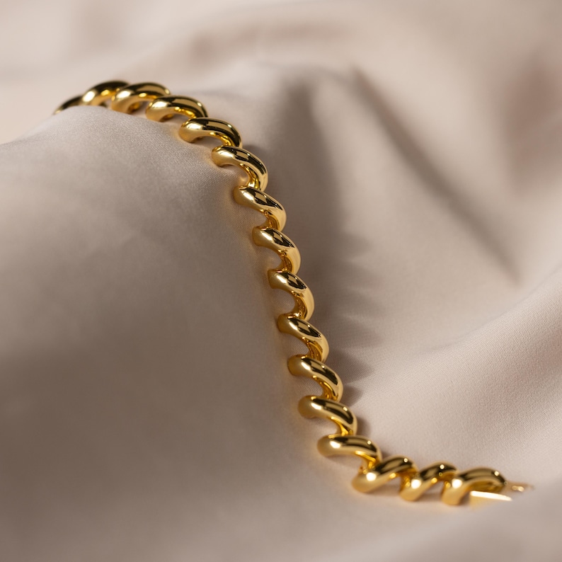 May include: A gold bracelet with a spiral design. The bracelet is made of linked, curved segments, creating a flowing, elegant look. The jewellery is displayed on a cream-coloured, textured fabric.
