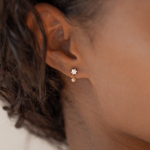 May include: Gold earrings with a small flower design and a dangling crystal.