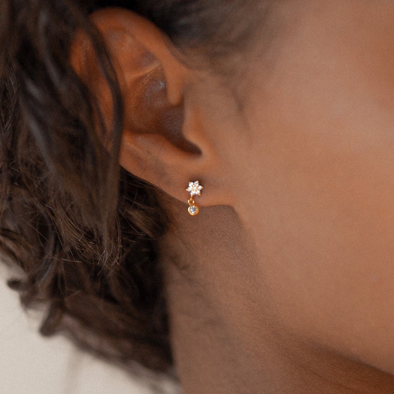 May include: Gold earrings with a small flower design and a dangling crystal.