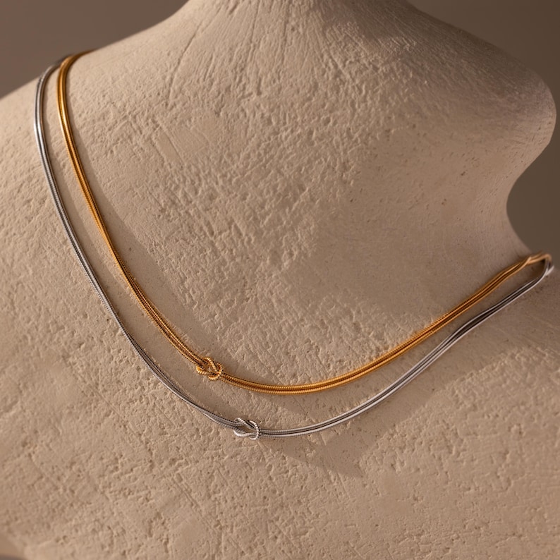 May include: Two delicate necklaces, one gold and one silver, displayed on a neutral-toned bust. Each necklace features a small knot detail. The gold necklace is positioned slightly above the silver one, creating a layered effect. Jewelry.