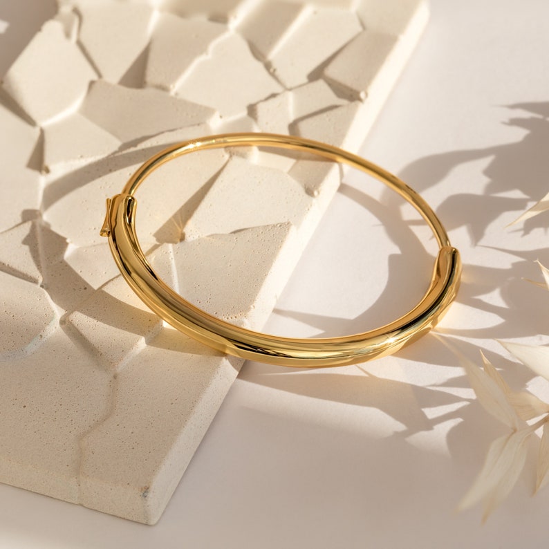 A close up of our Aris Hinge Bracelet in 18K gold placed on top of a white textured tile and white tabletop - featuring an about 73mm x 59mm and about 6mm thick statement bangle with a Hinge clasp design.