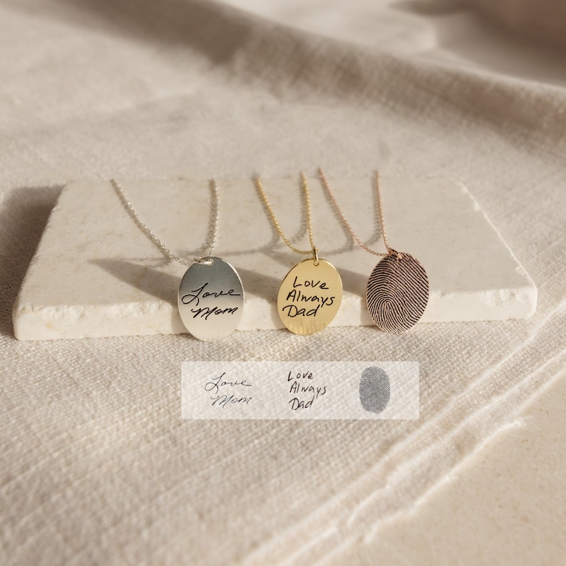 May include: Three silver, gold and rose gold necklaces with oval pendants. The silver pendant has a handwritten inscription that says "Love Mom". The gold pendant has an inscription that says "Love Always Dad". The rose gold pendant has a fingerprint design.
