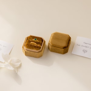 May include: Two gold rings with a green gemstone in a brown velvet ring box. The box is on a white surface with a white card that says "Happiness looks beautiful on you" with a heart design.