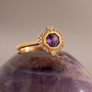 May include: A gold ring featuring a central hexagonal purple gemstone surrounded by a halo of small white round-cut diamonds. The setting is geometric, with two larger round diamonds positioned at the top and bottom of the hexagonal frame.