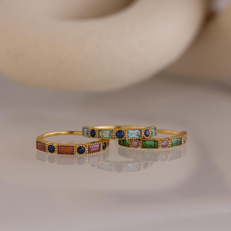 May include: Three gold-colored rings are displayed, each adorned with a series of rectangular and round gemstones in various colors. The rings feature a repeating pattern of gemstones, creating a colorful and elegant design.