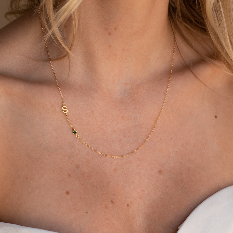 A close up of our Sideways Initial Birthstone Necklace in 18K Gold finish on our models neck hanging just below her collarbone - featuring a small 7mm letter S charm and a 3mm green emerald stone hanging sideways on a dainty cable chain.