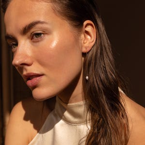 May include: A gold-toned threader earring featuring a single, small, white, teardrop-shaped pearl dangling from a thin, delicate metal chain. The earring is worn in the earlobe, with the chain extending downward from the piercing point.