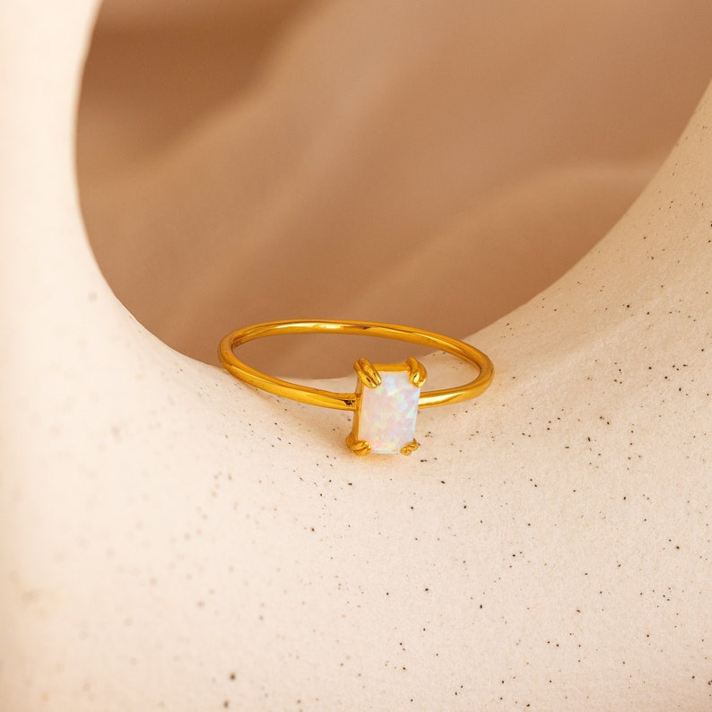 May include: A gold-colored ring with a rectangular, iridescent opal stone. The ring's band is thin and delicate, and the opal is set in a four-prong setting. The ring is displayed on a neutral-toned surface.