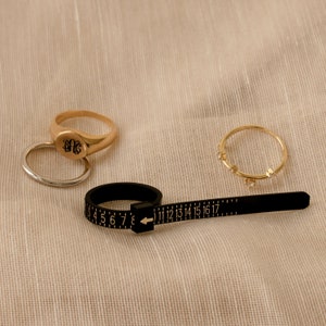 May include: A gold ring sizer with a black measuring tape and a gold ring with the initials "DG" engraved on it. There are two other gold rings on a beige fabric background.