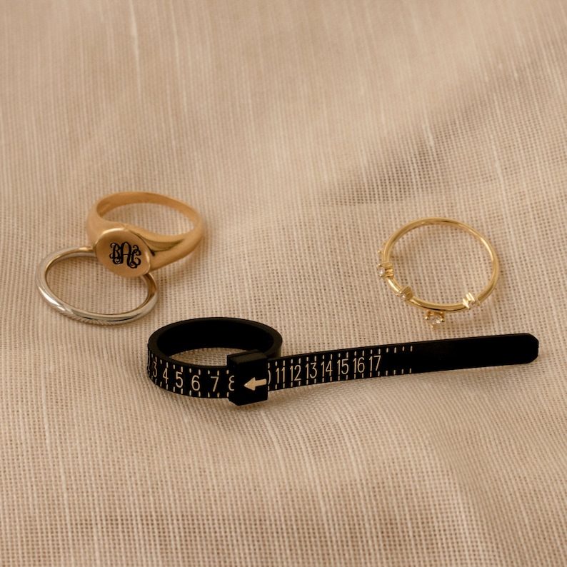 May include: A gold ring sizer with a black measuring tape and a gold ring with the initials "DG" engraved on it. There are two other gold rings on a beige fabric background.