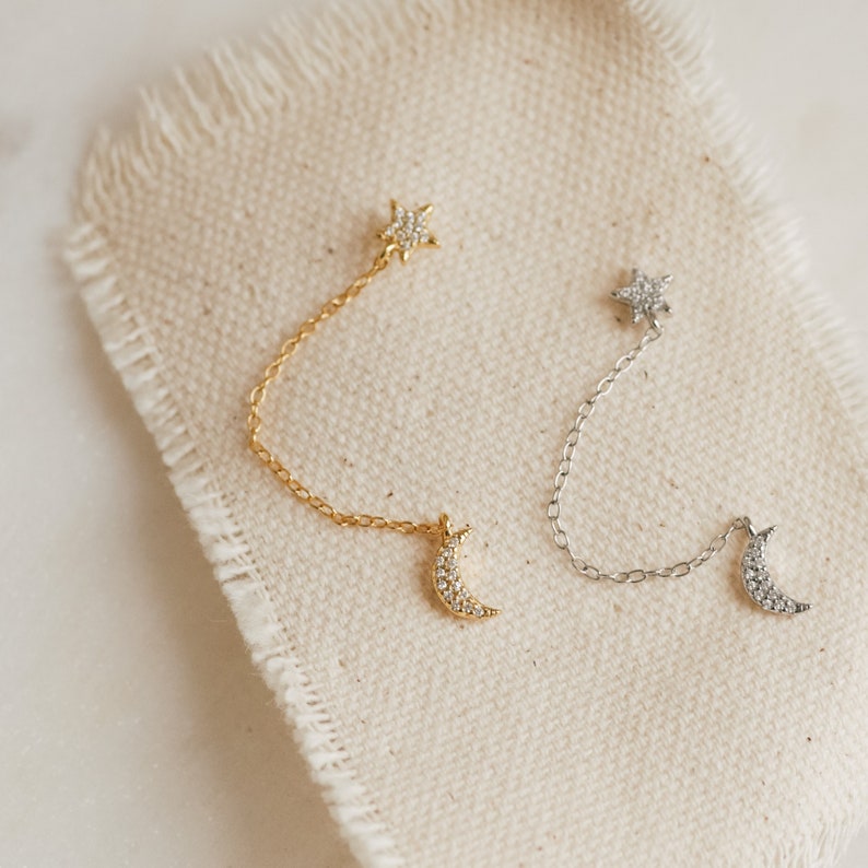 A gold earring and a silver earring lay flat on a beige piece of canvas. Both earrings have the same design; a pave star stud at the top and a pave moon stud at the bottom, connected by a dainty chain.